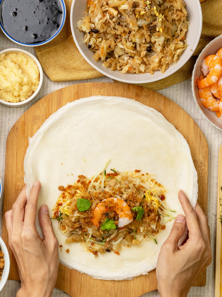 Hands preparing a golden popiah with fresh ingredients on a wooden board, showcasing culinary delicacy.