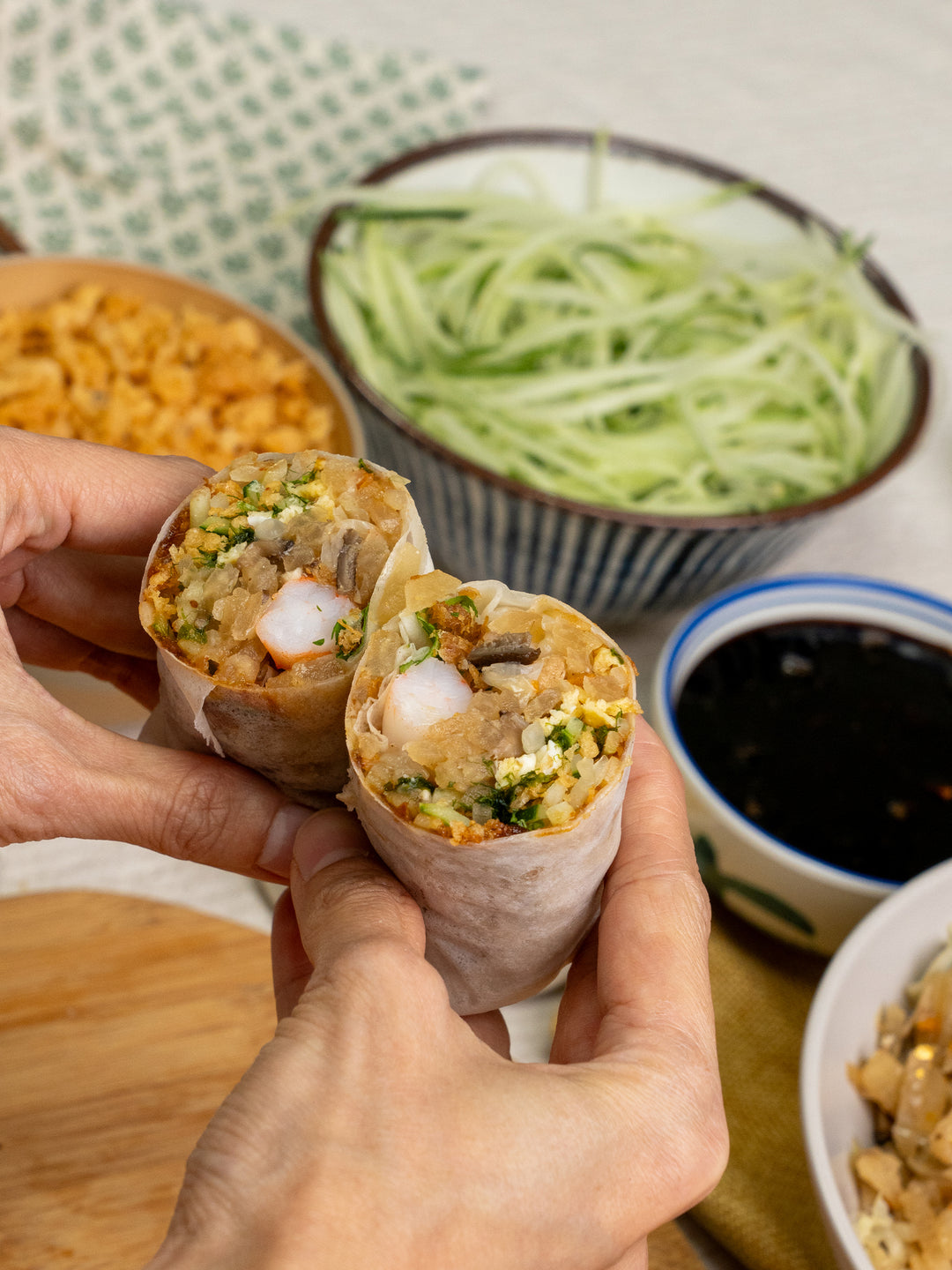 Two hands holding a freshly made golden popiah with shrimp, surrounded by ingredients like shredded cucumber and sauce.